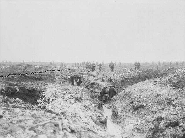 Black and white photograph. A photo of a large, muddy field with a Y shaped trench. Men, distant from the camera, walk through the trenches and look into dugouts. Beyond the trenches, men are visible in silhouette walking across the landscape.